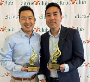 Two men in business-c casual attire pose with gold spiral trophies at an awards event, standing in front of a Citrus Club backdrop.
