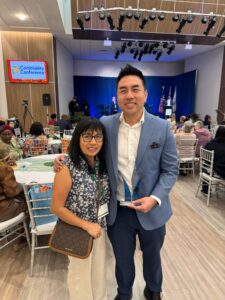 Smiling man in a blue suit and a woman with glasses pose for a selfie at a community conference banquet, awarding trophy in his hand.