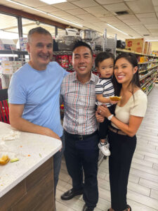 Smiling group of four in a grocery store bakery section, posing for a selfie with pastries on the counter behind them.
