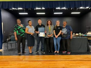 Group of seven adults standing on a stage, posing for a photo, one person holding a certificate folder and award ribbon in front.