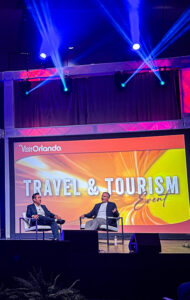 Two men in suits sit on a stage in a travel and tourism panel, with a bright backdrop reading 'Visit Orlando' and 'Travel & Tourism'.