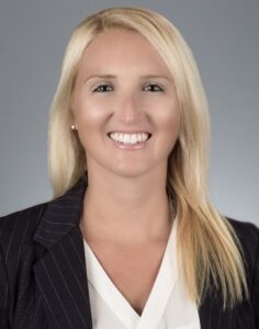 Professional headshot: smiling blonde woman in a dark pinstripe blazer against a gray background.