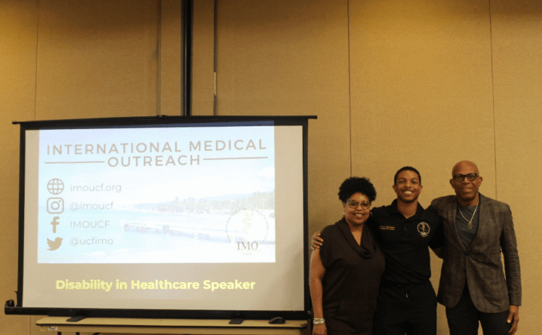 Three people pose together beside a projector screen displaying 'International Medical Outreach' and 'Disability in Healthcare Speaker'.