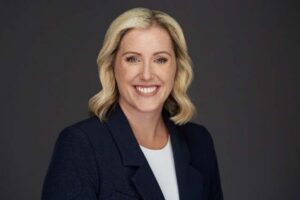 Portrait of a smiling professional woman with blonde hair, wearing a navy blazer and white blouse, posed against a dark background.