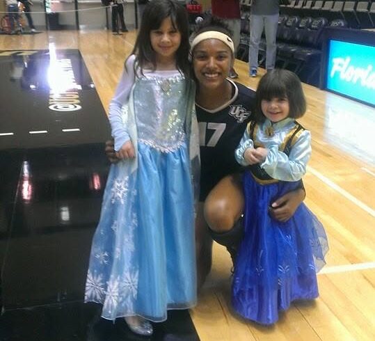 A female basketball player kneels on a gym floor with two young girls in blue princess dresses posing for a photo, with bleachers in the background.