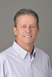 Portrait of a middle-aged man with short gray hair wearing a light checkered button-down shirt against a neutral gray studio background.