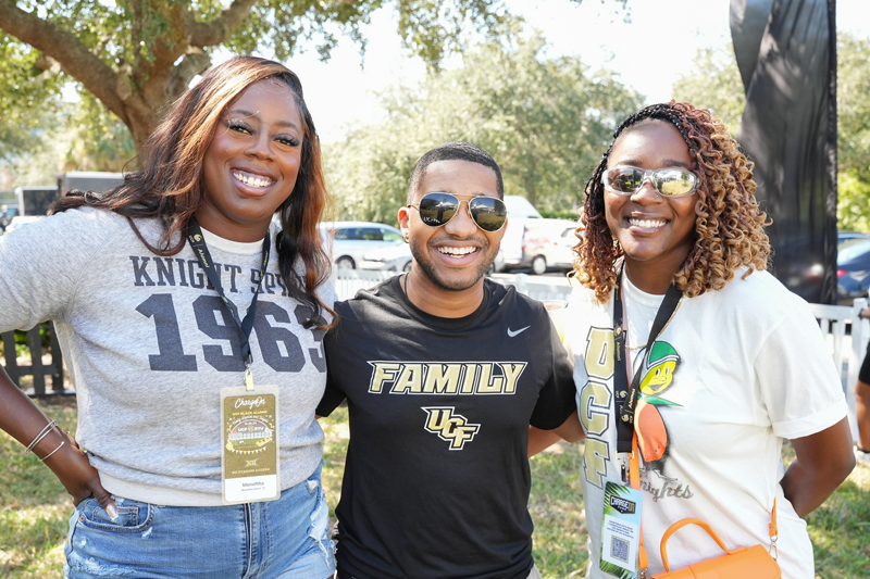 Three friends posing together at an outdoor event, all smiling and wearing lanyard badges.