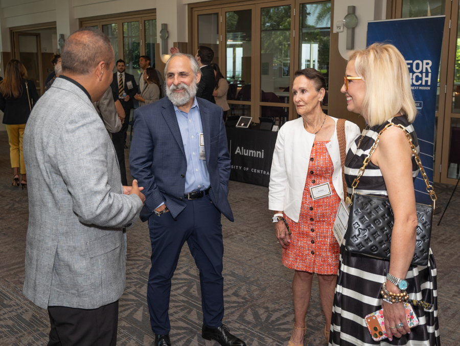 Four adults in business attire chat in a lobby at an alumni event, with a table labeled 'Alumni' and a blue banner nearby.