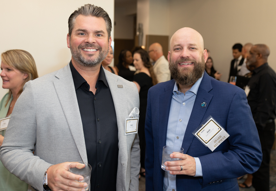 Two smiling men in suits at a social event, holding drinks with name badges on their jackets.