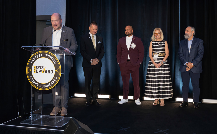Man speaks at a podium on stage while four formally dressed people stand in a line behind him.