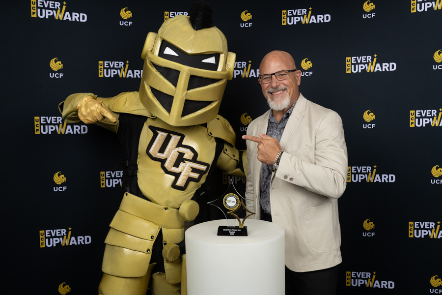 UCF mascot in gold armor posing with a smiling man in a light blazer beside a trophy on a white pedestal, against a branded backdrop.