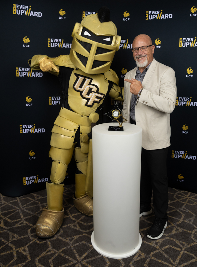 Gold armored mascot with 'UCF' on chest posing with a man at an award ceremony, trophy on a pedestal in front of a branded backdrop.
