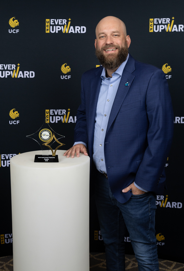 Man in a blue blazer and light shirt stands next to a white pedestal with an award trophy, at an event backdrop reading 'Ever Upward' and UCF logos.