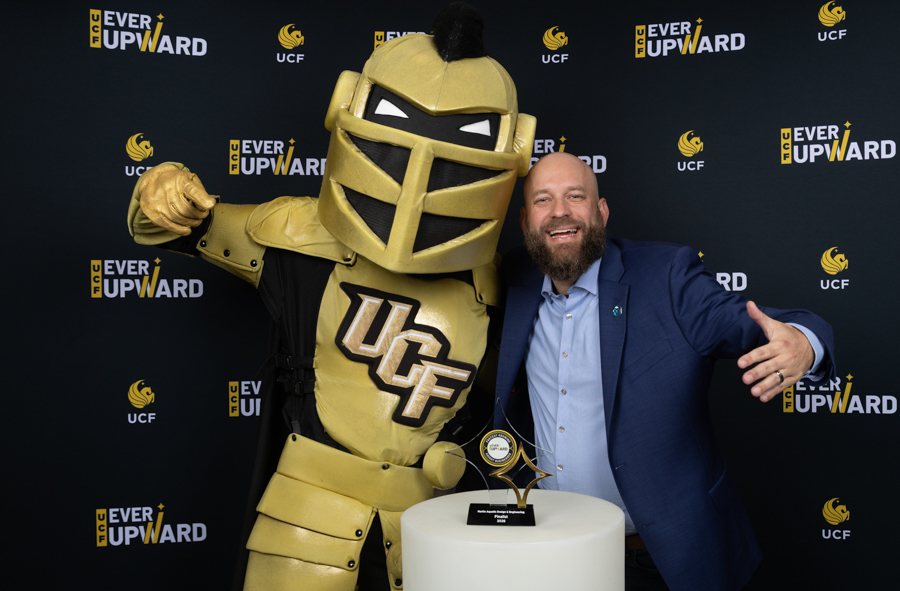 Golden UCF Knights mascot with a man in a blue suit posing beside a trophy on a black backdrop with Ever Upward and UCF logos.