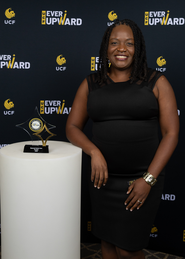 Woman in a black dress smiles beside a trophy on a white pedestal at a UCF 'Ever Upward' backdrop