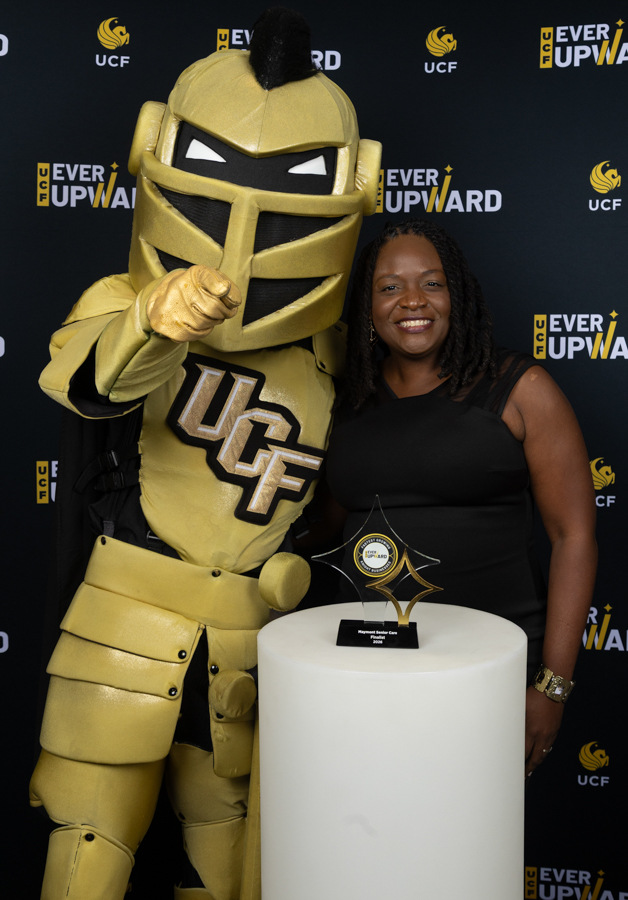UCF golden knight mascot posing with a smiling woman beside a white pedestal trophy at a 'Ever Upward' event backdrop.
