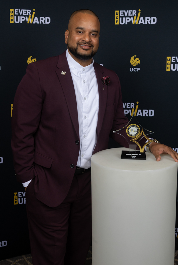 Man in a burgundy suit and white shirt smiles beside a star-shaped award on a white pedestal at a UCF Ever Upward event backdrop.