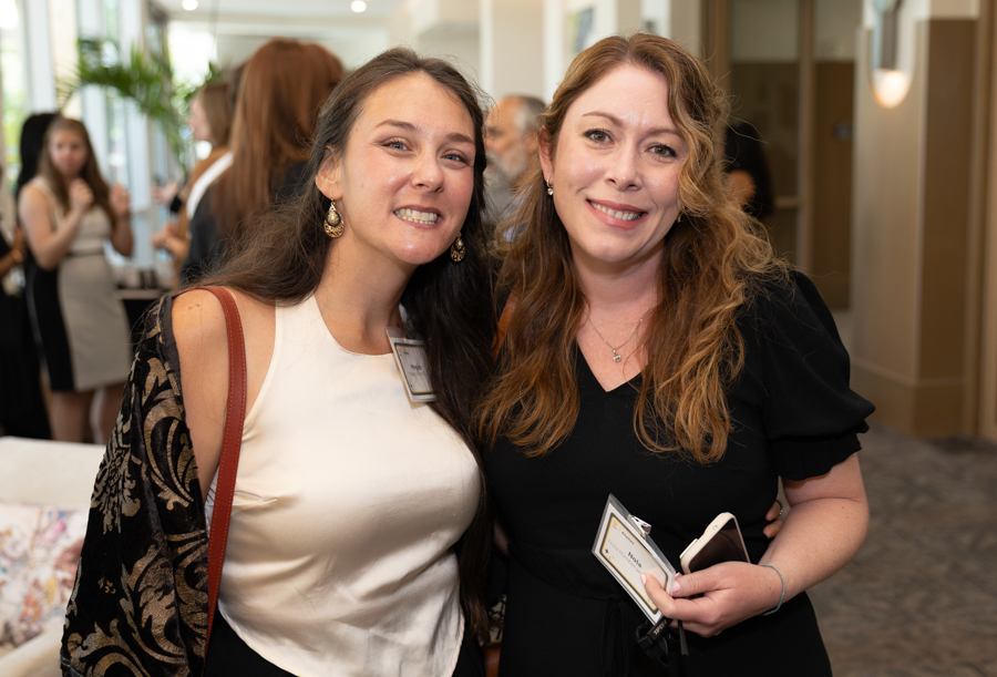 Two women at a social event pose for a photo, both wearing name badges and smiling.