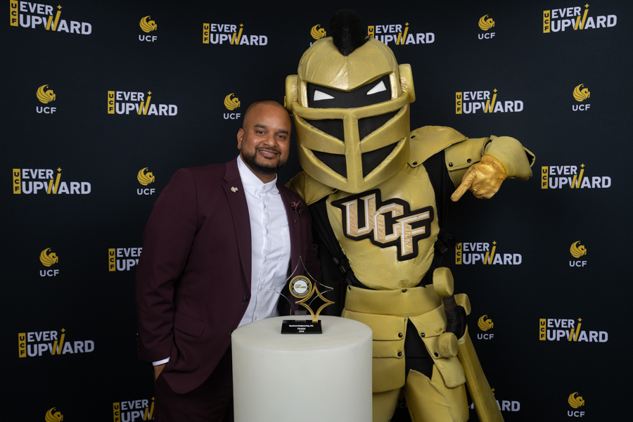 Man in a burgundy suit poses with the UCF Golden Knight mascot at an Ever Upward event, beside a white pedestal trophy.
