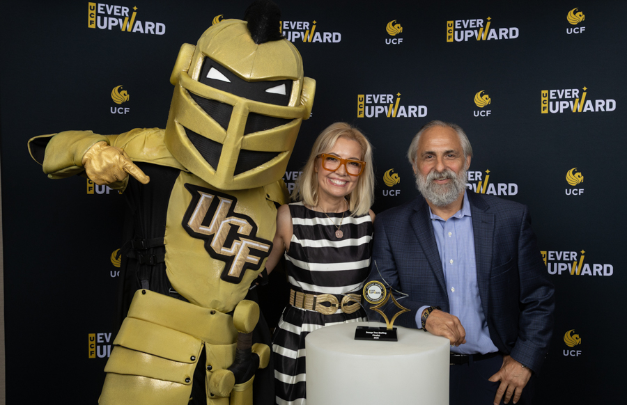 Gold-armored UCF mascot poses with a smiling blonde woman in a striped dress and a bearded man in a blue blazer; a trophy rests on a pedestal as they stand at an 'Ever Upward' event backdrop.