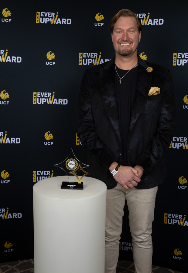 Smiling man in a black velvet blazer and beige pants stands beside a star-shaped trophy on a white pedestal at a UCF 'Ever Upward' event.