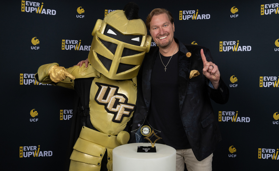 Man in a black shirt and blazer poses with a gold UCF Knight mascot and a trophy on a white pedestal at an Ever Upward event.