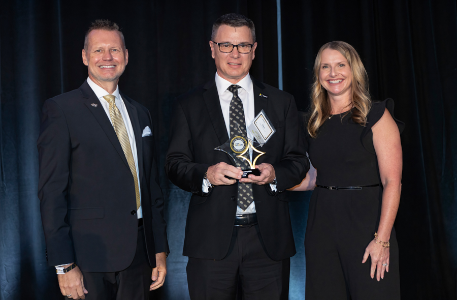 Three people in formal attire pose on stage; center man holds an award trophy, flanked by a man and a woman on a dark backdrop.