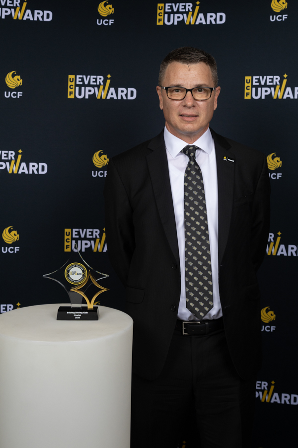 Man in a black suit and glasses standing beside a star-shaped trophy on a white pedestal at a UCF Ever Upward event backdrop.