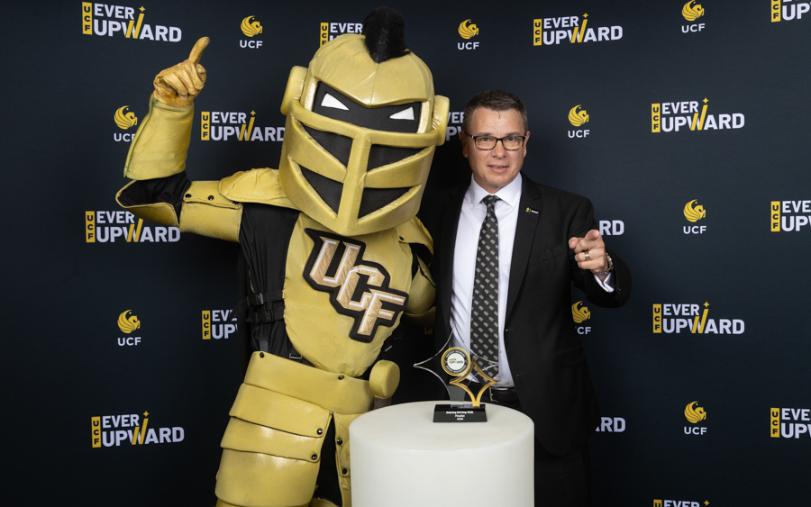 UCF Golden Knight mascot and a man in a suit pose with a trophy at an 'Ever Upward' event backdrop.