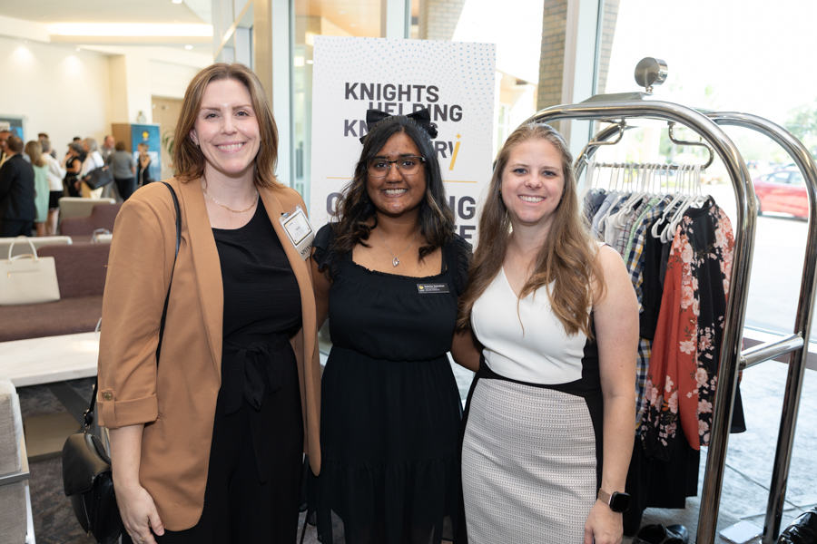 Three smiling women posing together at an indoor event, with a 'Knights Helping Knights' banner behind them.