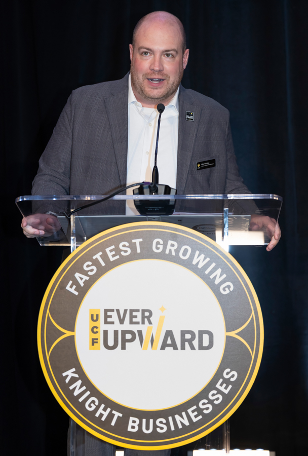 Man in a gray blazer speaks at a podium, with a circular event sign reading 'FASTEST GROWING KNIGHT BUSINESSES' and 'UCF UPWARD' in the center.