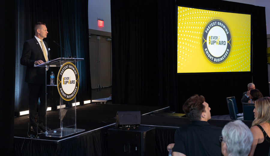 Male presenter in a suit stands at a podium with an Ever Upward logo, addressing a conference audience on a stage.