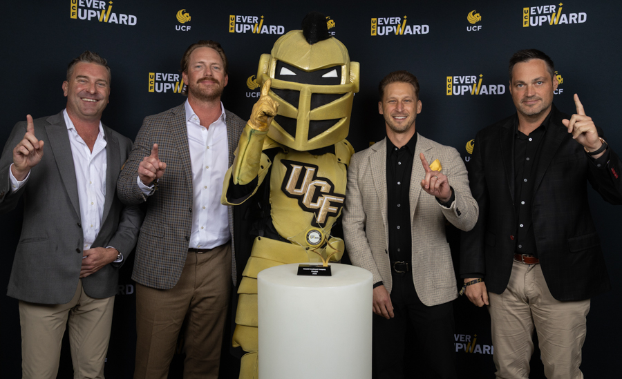 Five people pose with a gold Knight mascot and trophy on a pedestal at a UCF 'Ever Upward' event backdrop.