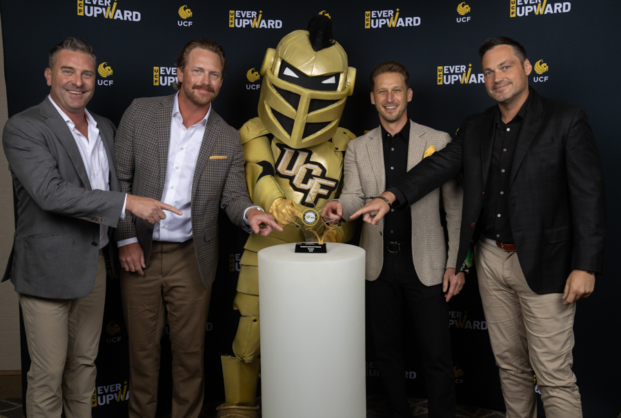 Four men in suits pose with a gold knight mascot, pointing to a trophy on a pedestal at a UCF 'Ever Upward' event.