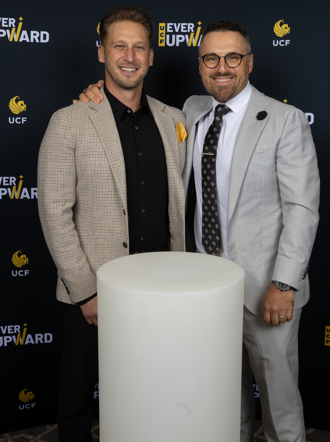 Two men in suits smiling for the camera at an event, one with an arm around the other, standing beside a white pedestal with a dark backdrop.