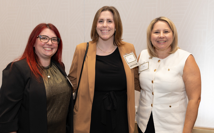 Three women in business attire posing together at an event, smiling for the camera.