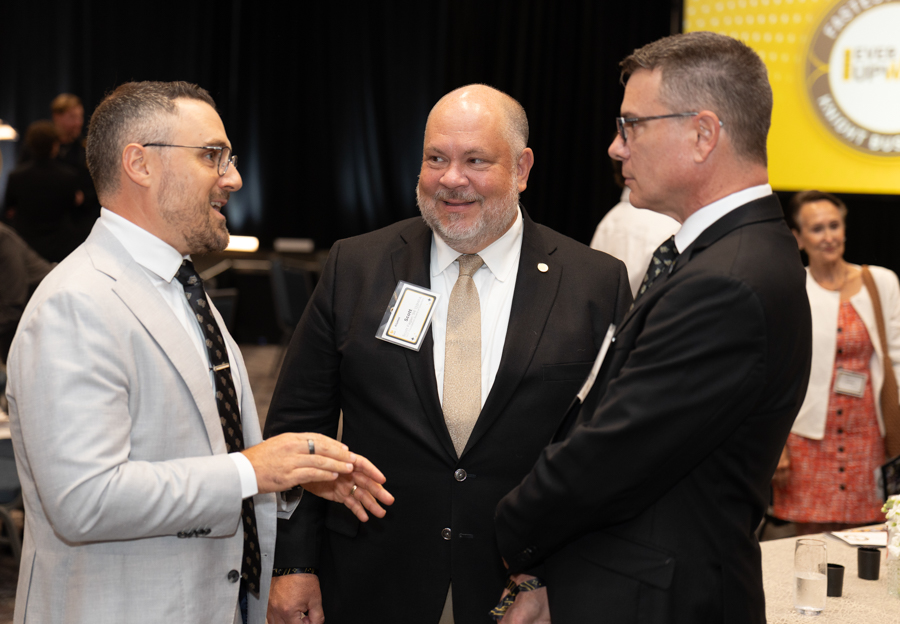 Three men in suits talk and smile at a conference, badges visible, with a yellow display in the background.