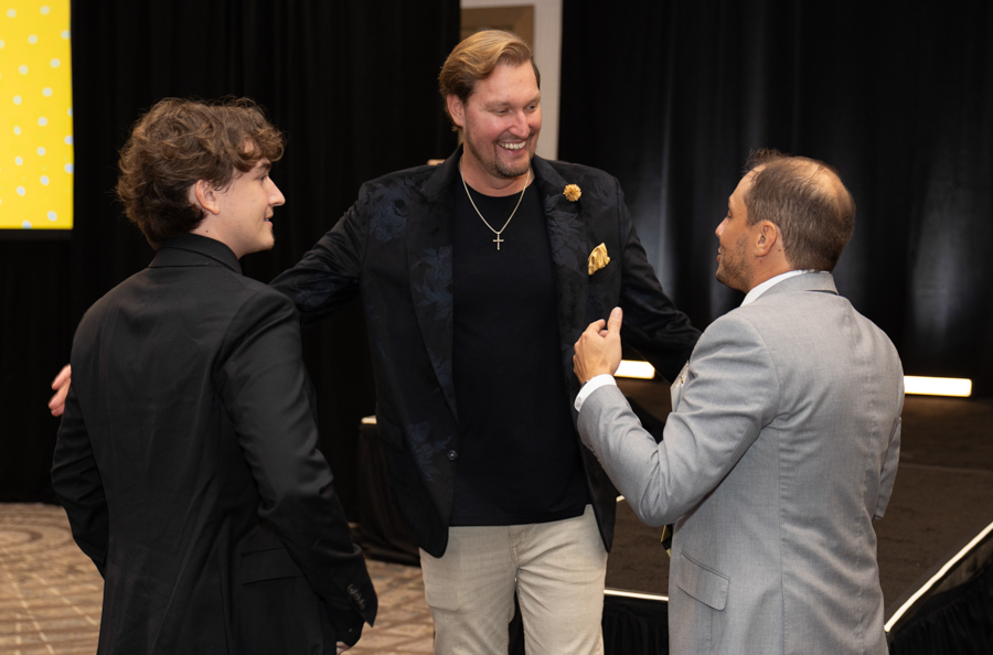 Three men in suits converse on a backstage event area; the center man wears a black blazer, cross necklace, and beige pants.