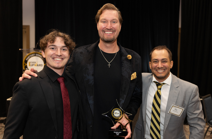 Three men in suits smile for a photo at an awards event, with one man holding a star-shaped trophy and a badge visible on another.