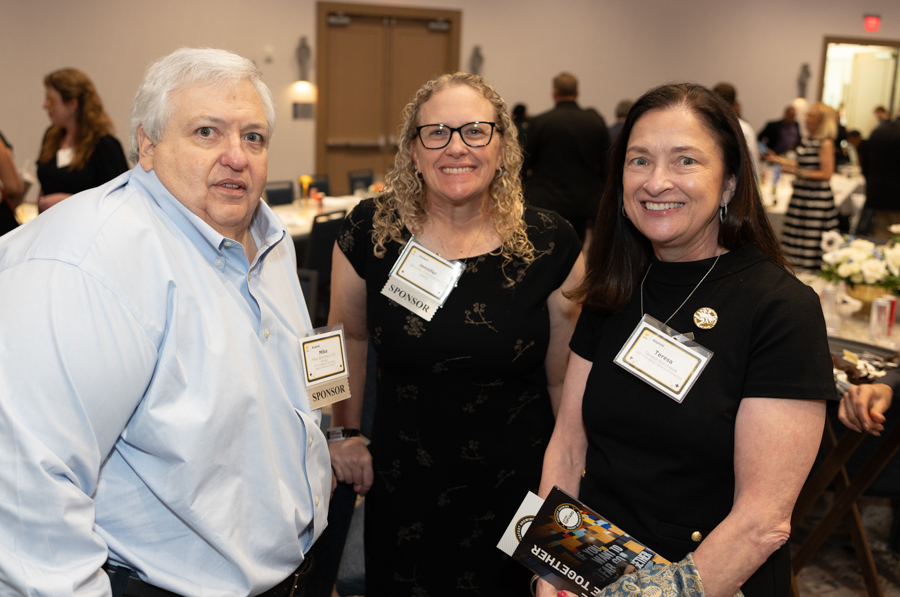 Three smiling attendees wearing sponsor badges pose for a photo at a formal event in a banquet hall, with tables in the background.