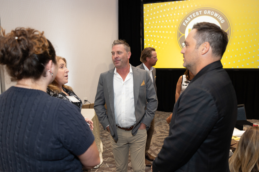 Group of professionals in business attire talking in a conference room, with a bright yellow stage backdrop reading 'FASTEST GROWING'.