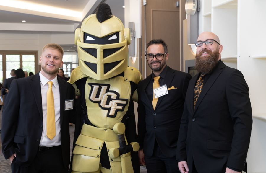 Three men in suits posing with a gold knight mascot in a hallway at an event outside UC letters on chest visible (UCF).