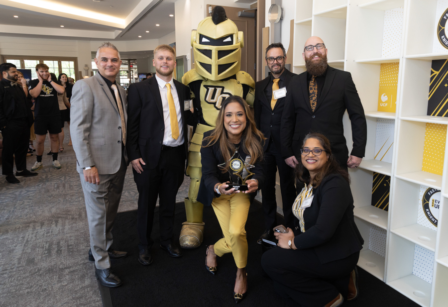 Group of professionals posing with a gold knight mascot; a woman in front holds a trophy at an awards event.