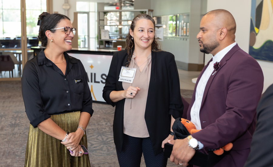 Three professionals chat and laugh in a bright lobby: two women in business attire and a man in a burgundy suit, one wearing a sponsor badge and holding an orange plush toy.