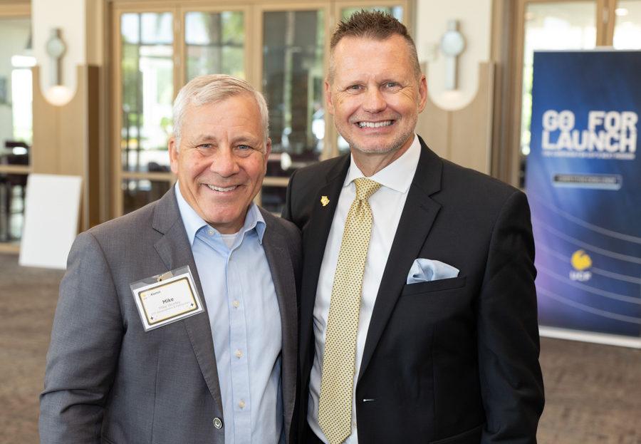 Two smiling men in business attire pose together at a conference lobby, one wearing a name badge on his gray suit.