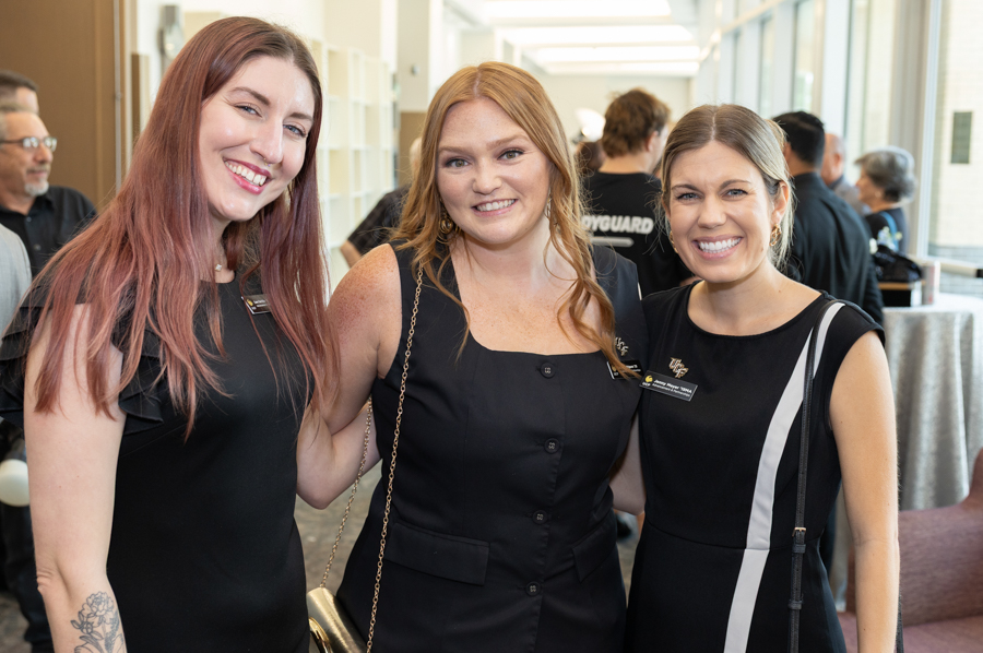 Three women in black dresses pose and smile at the camera at an indoor networking event.