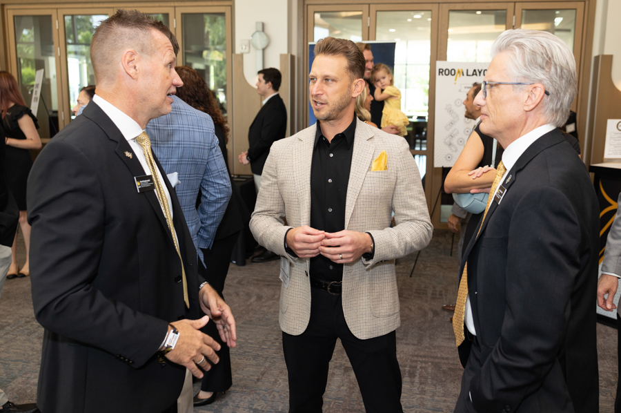 Three men in business attire converse at a networking event, with attendees and display banners in a conference room.