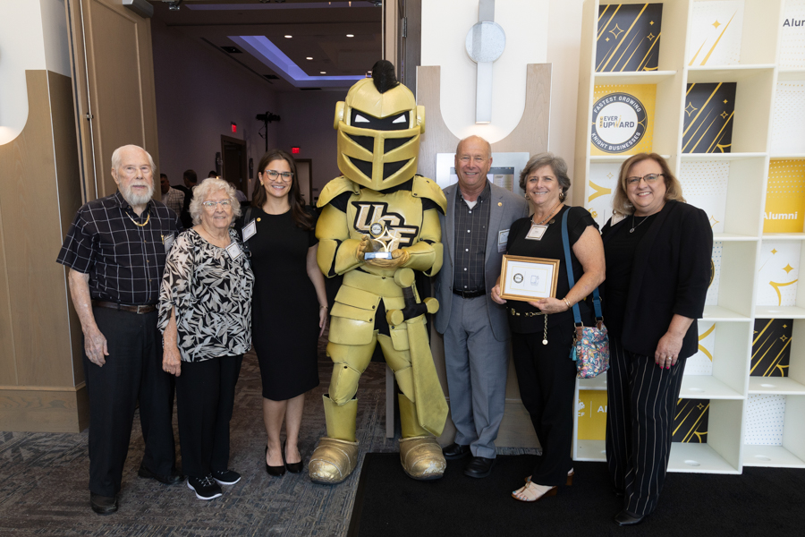 Seven people posing for a group photo with a large gold knight mascot inside a campus lobby; one person holds a framed certificate.