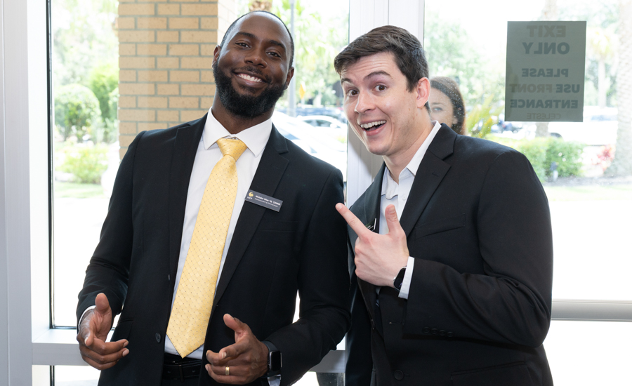 Two men in suits pose for a friendly photo at an office entrance; one wears a yellow tie and name badge, smiling at the camera with the other man pointing playfully.