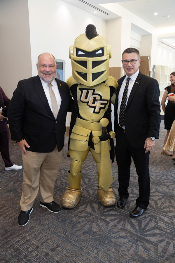 Two men in suits pose with a gold UCF Knights mascot in a hallway lobby.